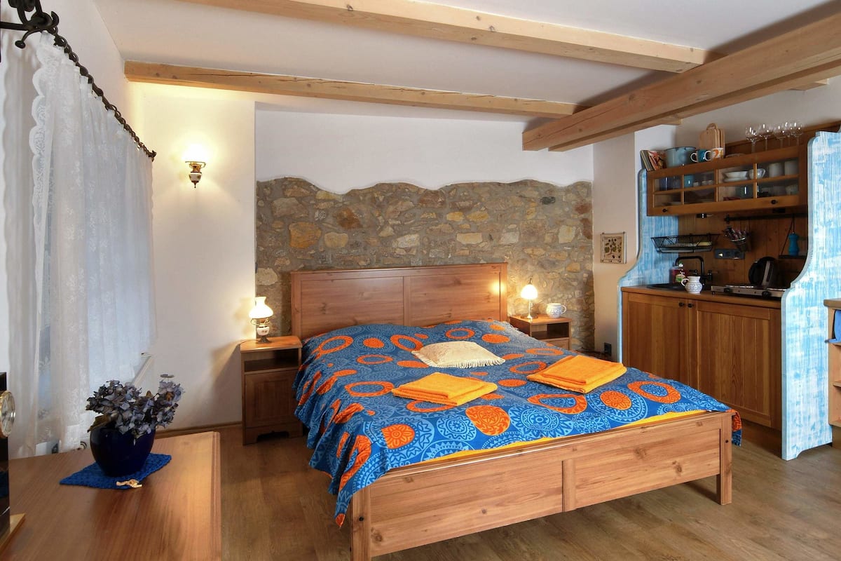 A bedroom with a wooden bed, a stone wall, and a kitchenette.