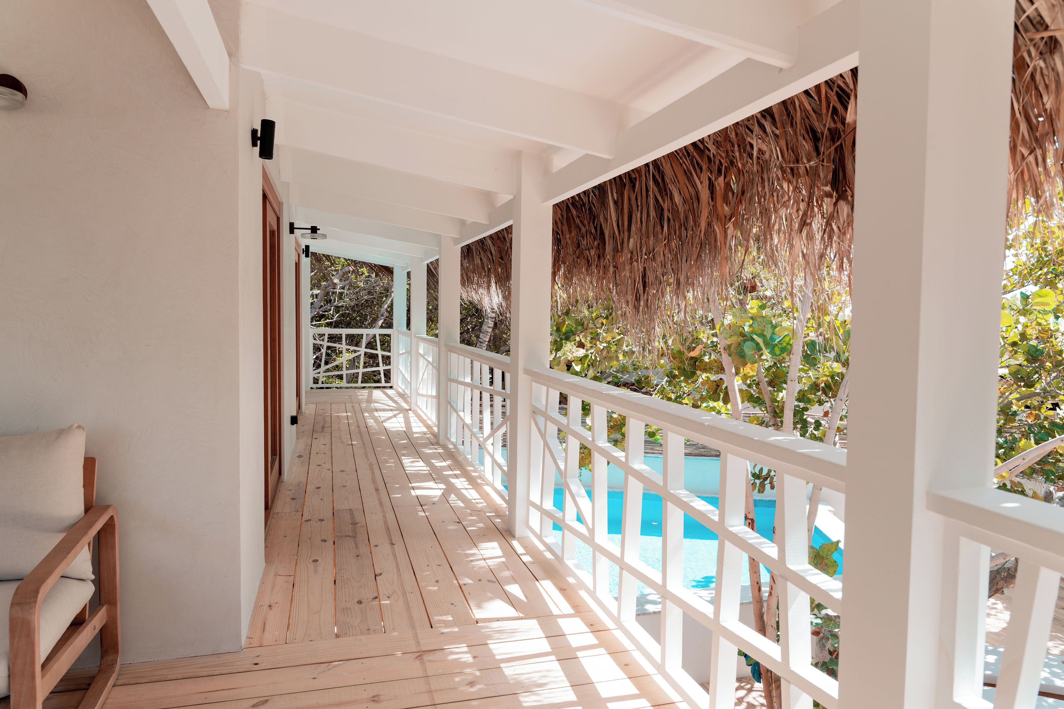 A wooden deck with a white railing, leading to a pool area with a thatched roof and greenery.
