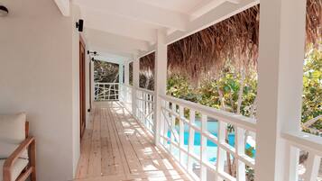 A wooden deck with a white railing, leading to a pool area with a thatched roof and greenery.