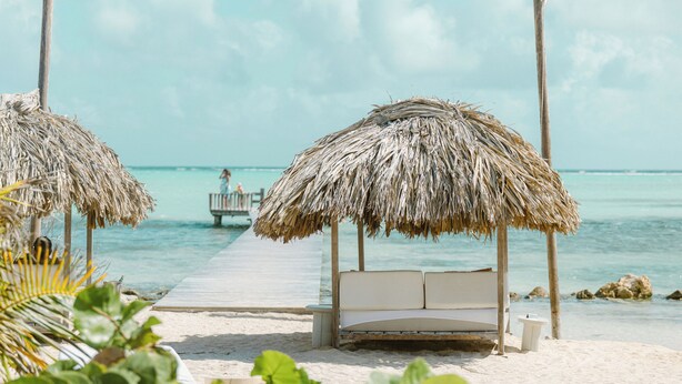 Una playa con sombrillas de paja y una terraza de madera que conduce a una cama blanca con vista al océano.
