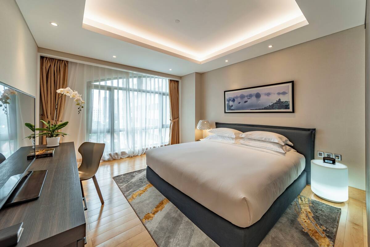 Een moderne hotelkamer met een groot bed, een bureau en een stoel. Er is een raam met gordijnen, een schilderij aan de muur en een lamp op het nachtkastje.
