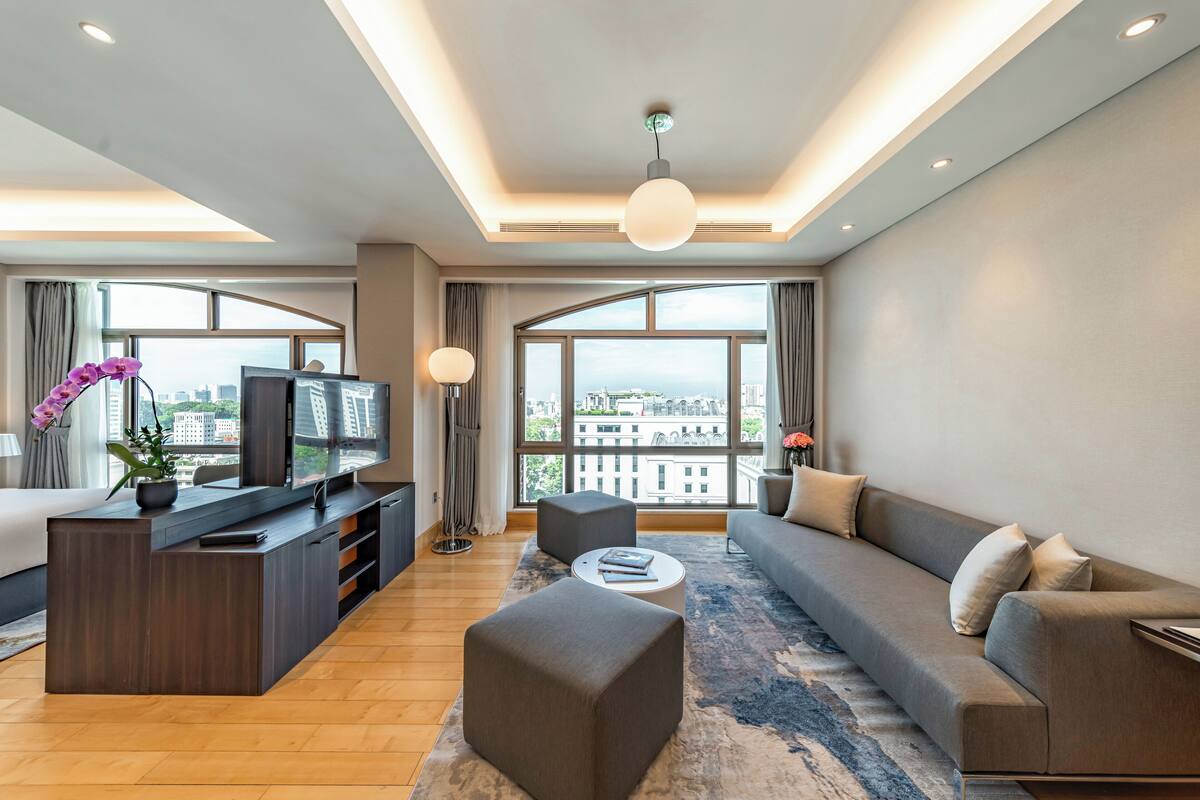 Een moderne hotelkamer met een bank, voetenbankje, televisie en grote ramen met uitzicht op de stad.