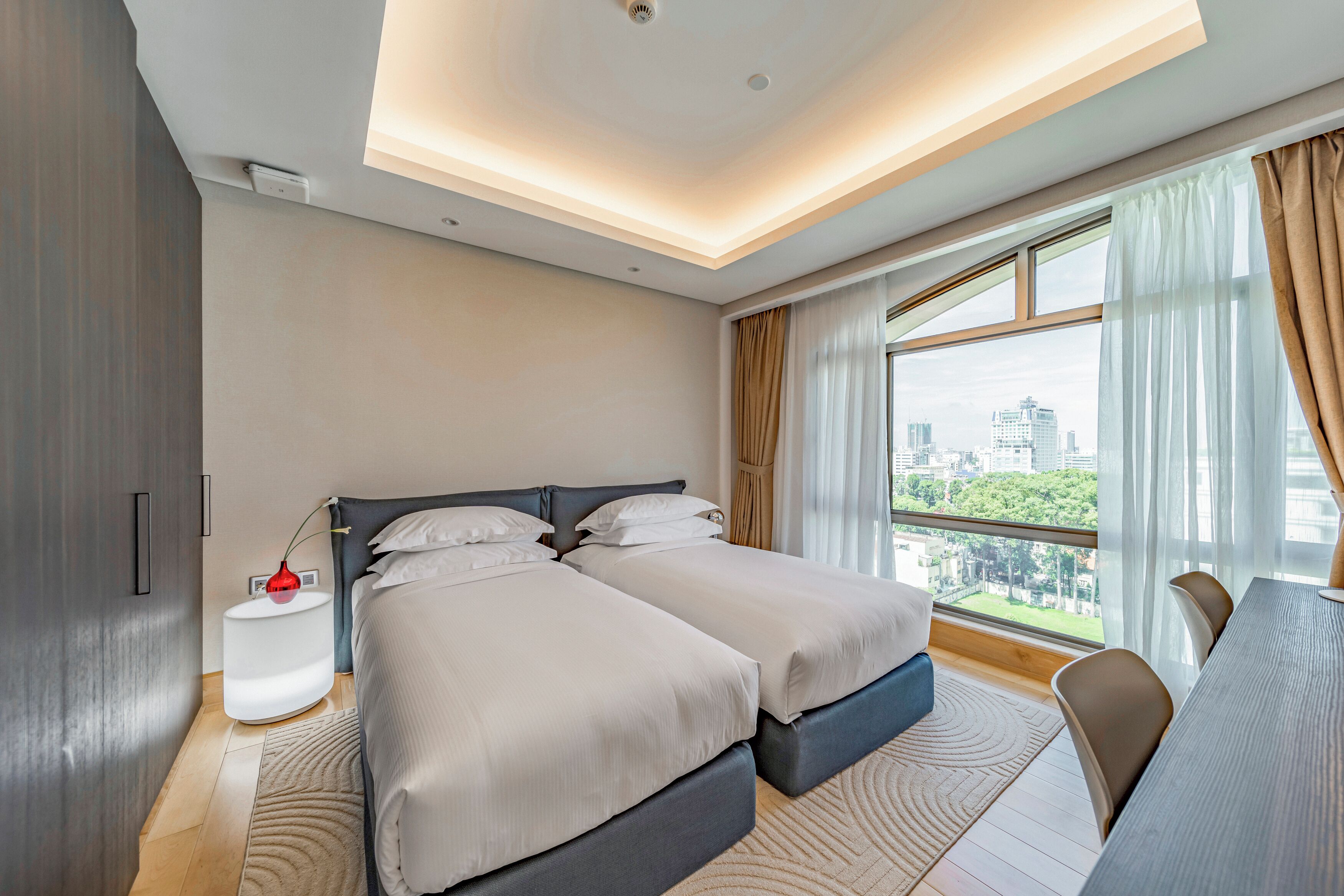 A modern hotel room with two beds, a dining table, and a large window with a city view.