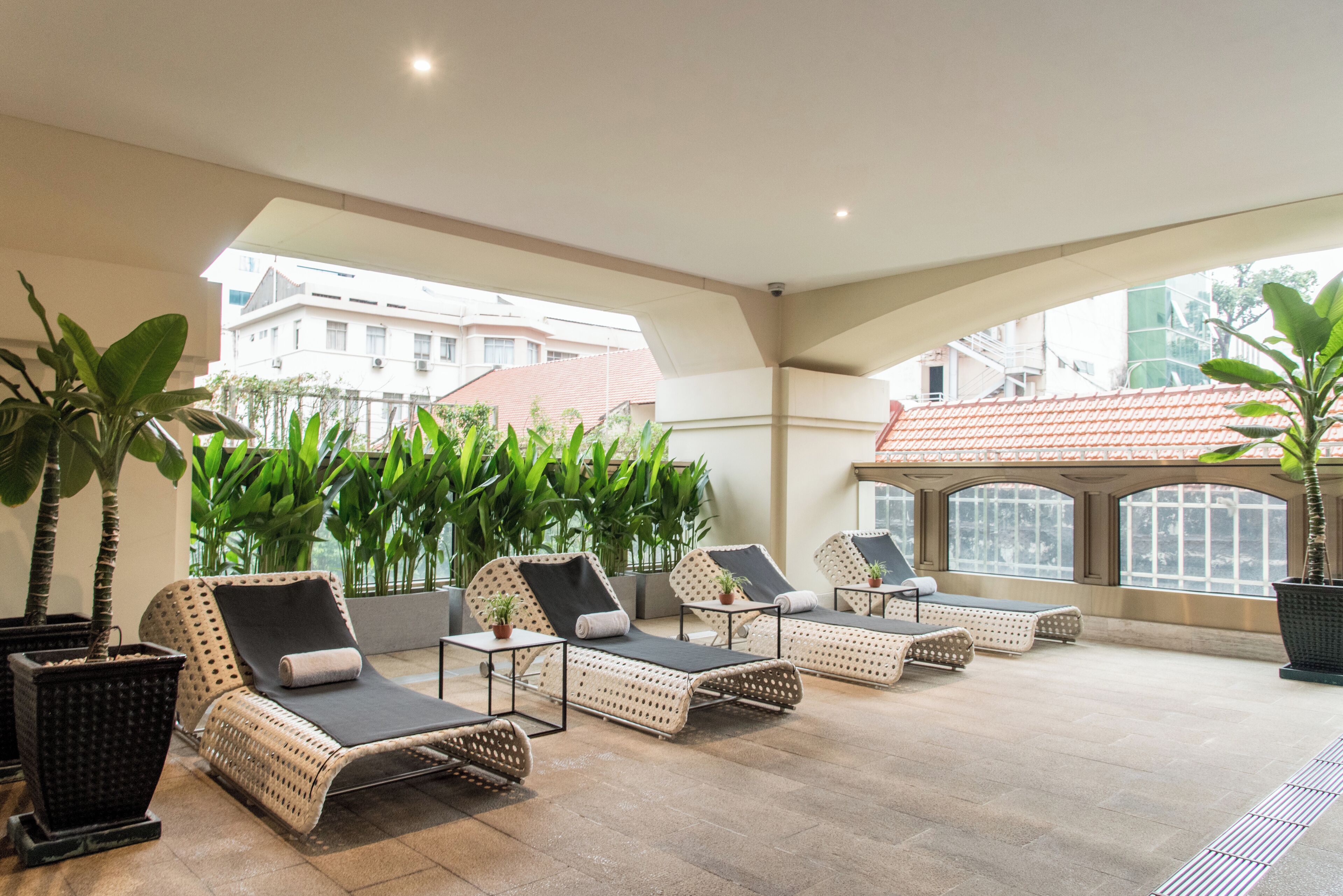 A sunlit outdoor lounge area with wicker lounge chairs, potted plants, and a view of buildings in the background.