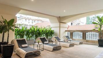 A sunlit outdoor lounge area with wicker lounge chairs, potted plants, and a view of buildings in the background.