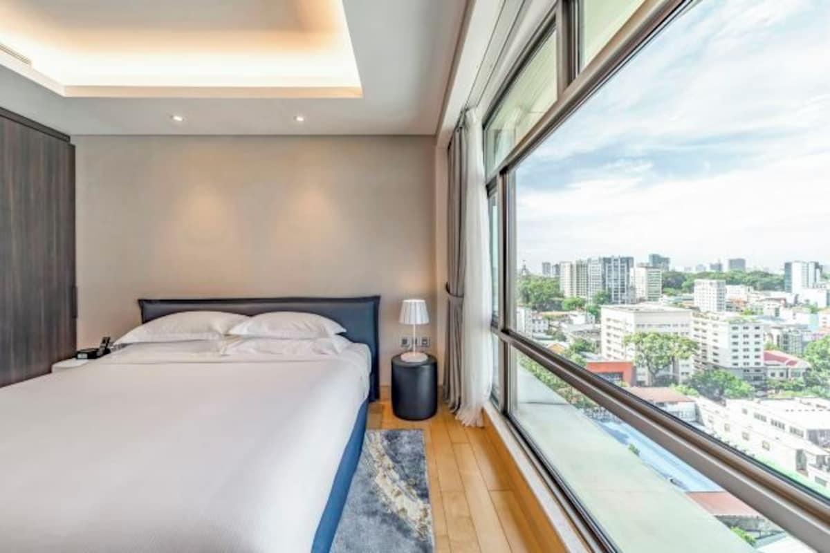 Ett modernt hotellrum med en stor säng, ett sängbord, en lampa och utsikt över staden genom stora fönster.