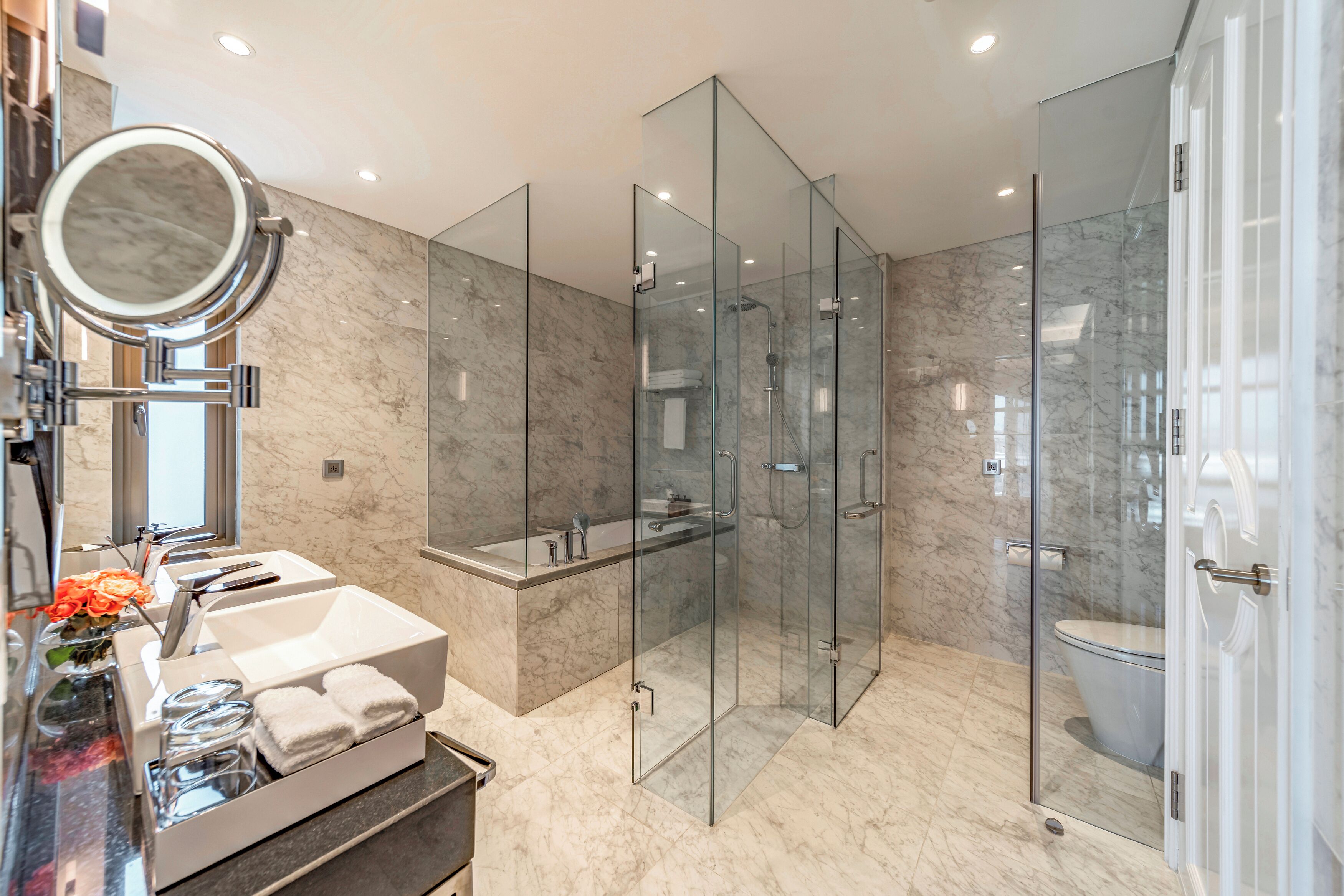 Un baño moderno con ducha envuelta en vidrio, suelo de mármol, tocador con lavamanos y espejo, y un inodoro.