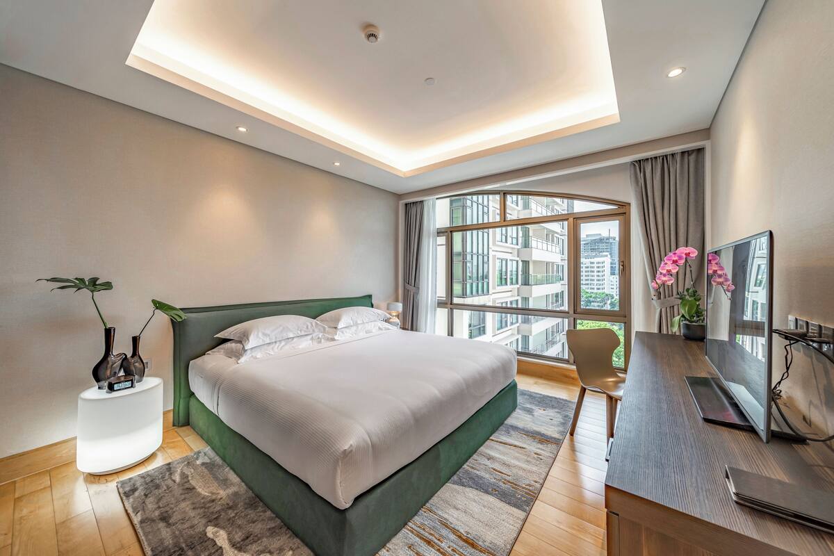 Een moderne slaapkamer met een groot bed, een bureau met een computer en stadszicht door grote ramen.