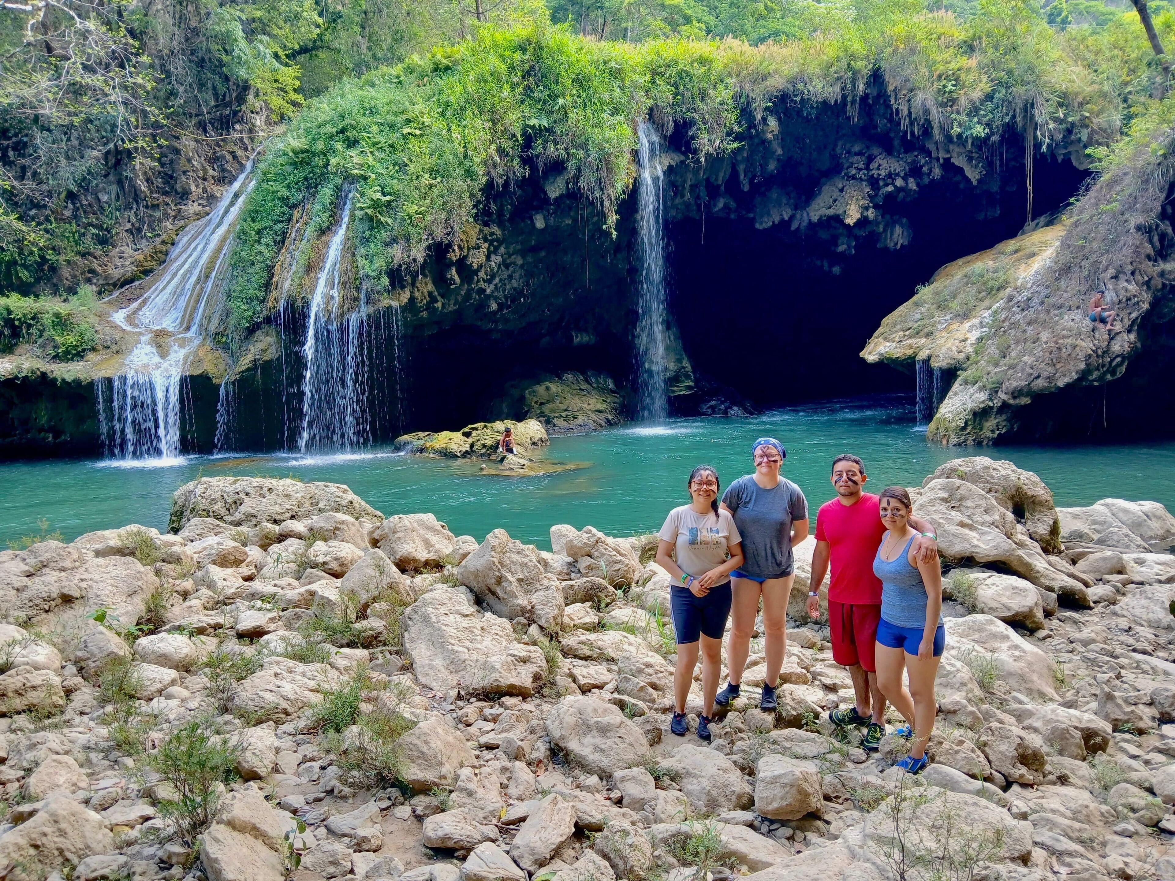Eine Gruppe von Menschen steht auf felsigem Gelände, im Hintergrund ein Wasserfall und ein klares, blaues Becken.