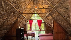 Una habitación con techo de paja, paredes de madera y una ventana con cortinas rojas. Se observan camas y una mesa con sillas.