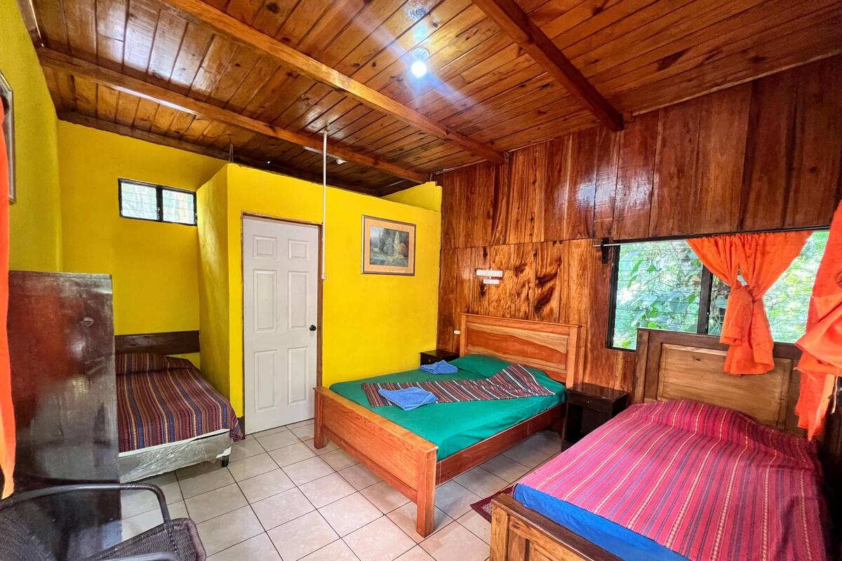 Una habitación con techos de madera, una cama con colcha verde, una silla y una pequeña mesa.