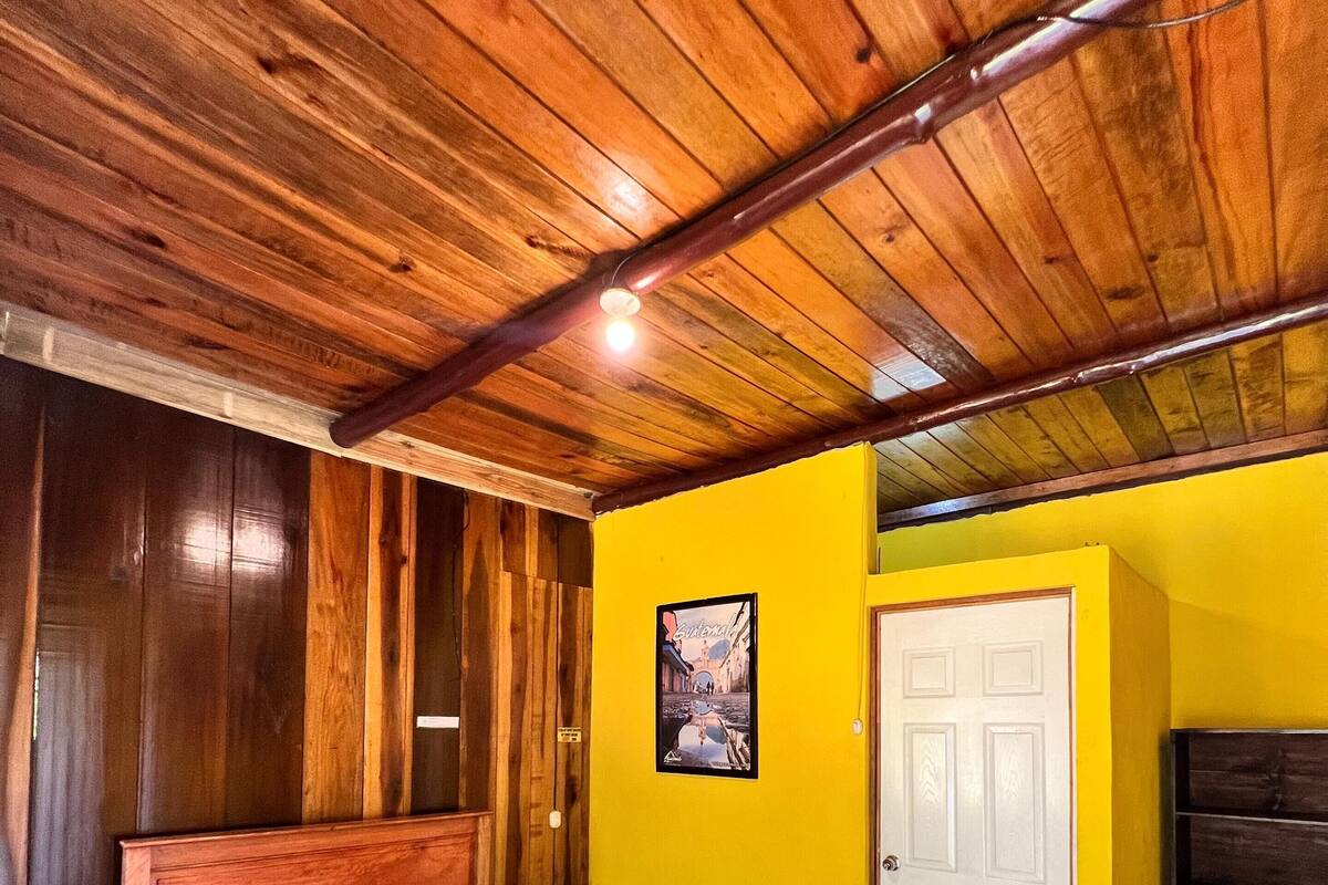 Techo de madera, una cama con colcha colorida, una puerta blanca y una pequeña mesa con mantel a rayas.