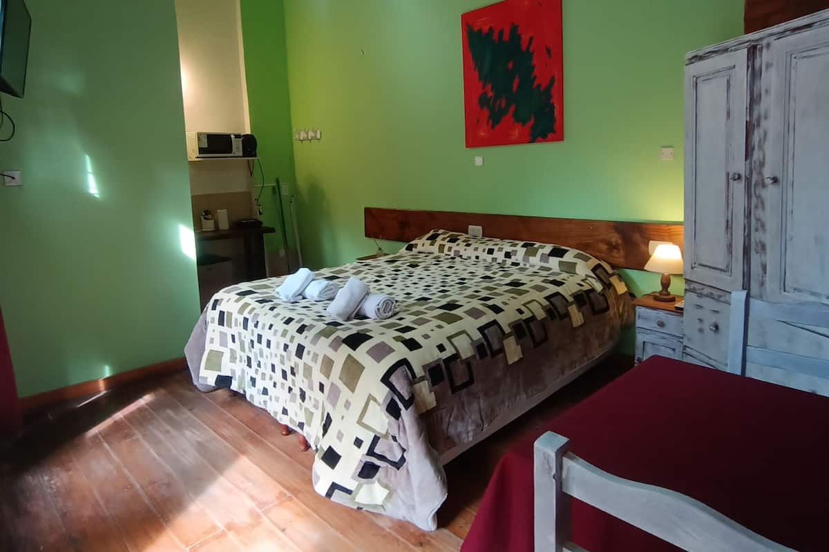 Una habitación con cama, armario de madera, una silla roja, una obra de arte en la pared y una lámpara colgante.