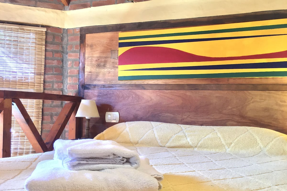 Una habitación acogedora con una cama, techo de madera, pared de ladrillo y un cuadro abstracto y colorido.