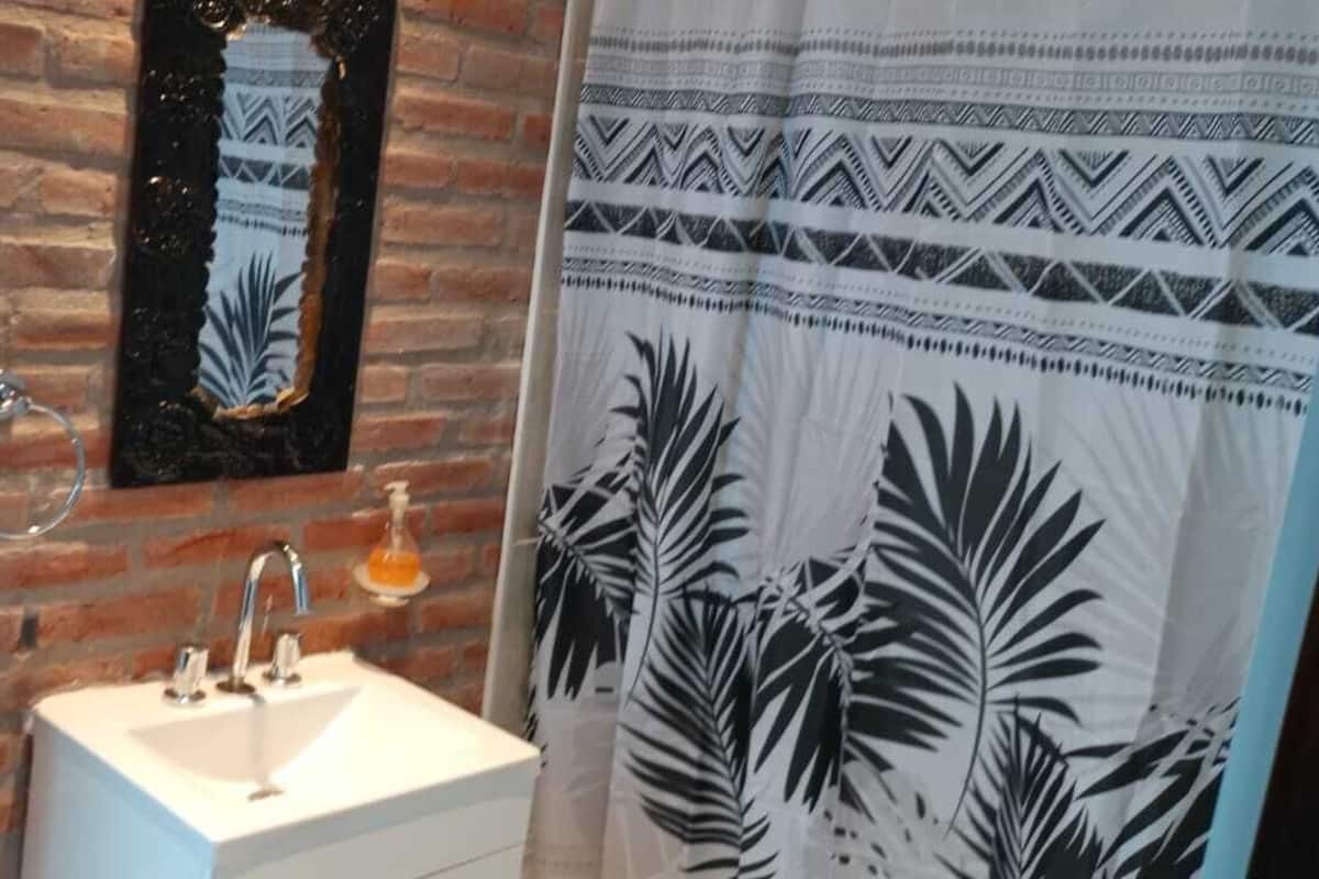 Un baño con lavamanos blanco, un espejo y cortina de ducha con estampado de hojas tropicales.