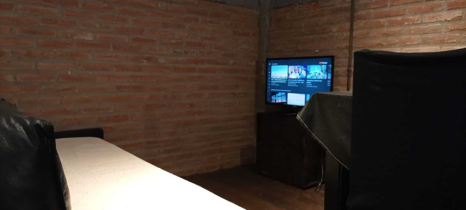 Une pièce avec un mur en briques, un téléviseur posé sur un meuble et un lit recouvert d’un drap blanc.