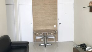Una habitación moderna con una mesa de comedor de vidrio, dos puertas blancas, un sofá negro y un pequeño estante de madera con un objeto decorativo.