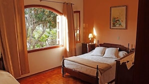 A bedroom with a wooden bed, a nightstand, a lamp, a window with curtains, and a painting on the wall. - Pousada Amanhecer na Mantiqueira (Águas de Lindóia)