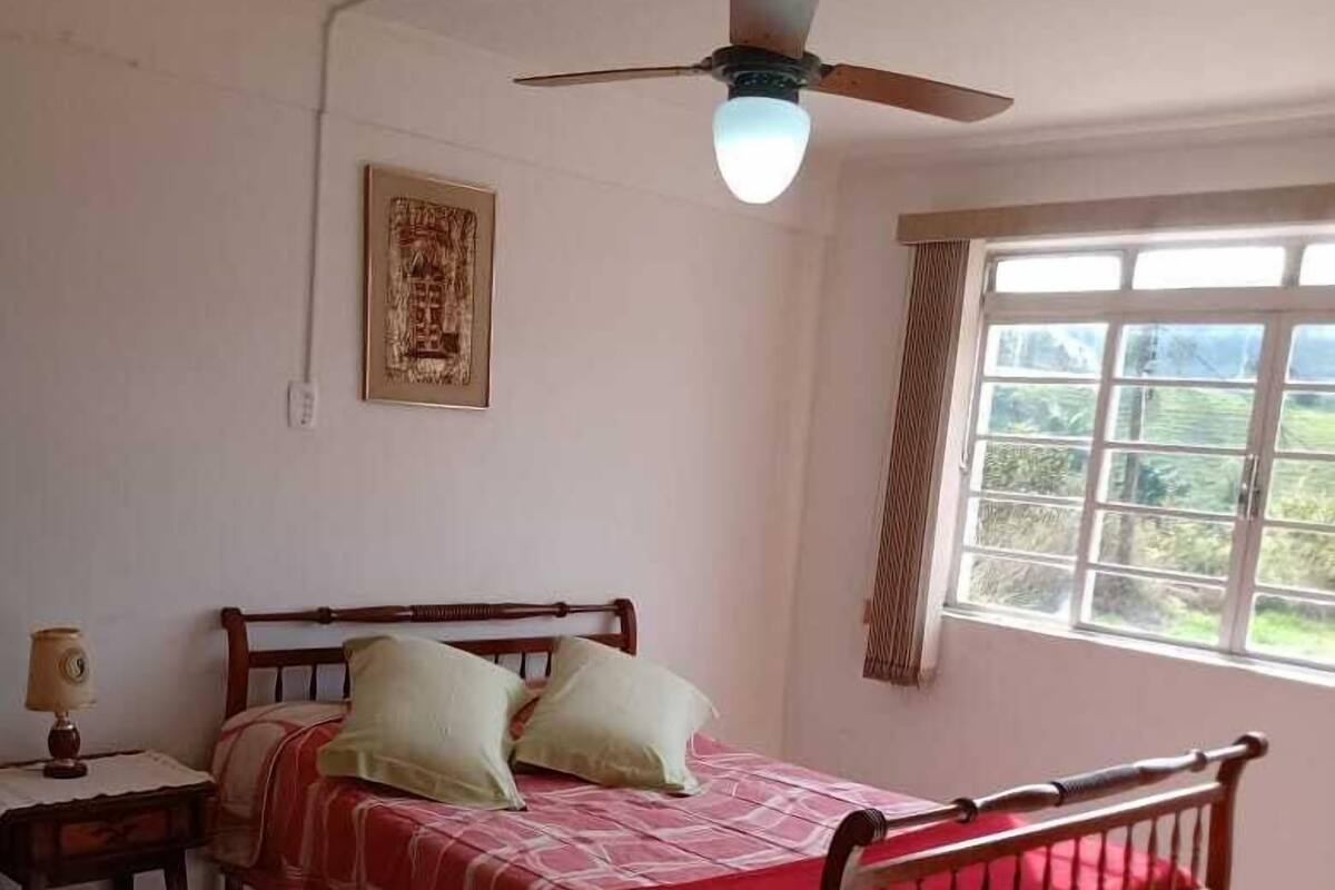 Ein Schlafzimmer mit einem Bett, einem Nachttisch, einem Deckenventilator, einem Fenster mit Vorhängen und einem gerahmten Bild an der Wand.