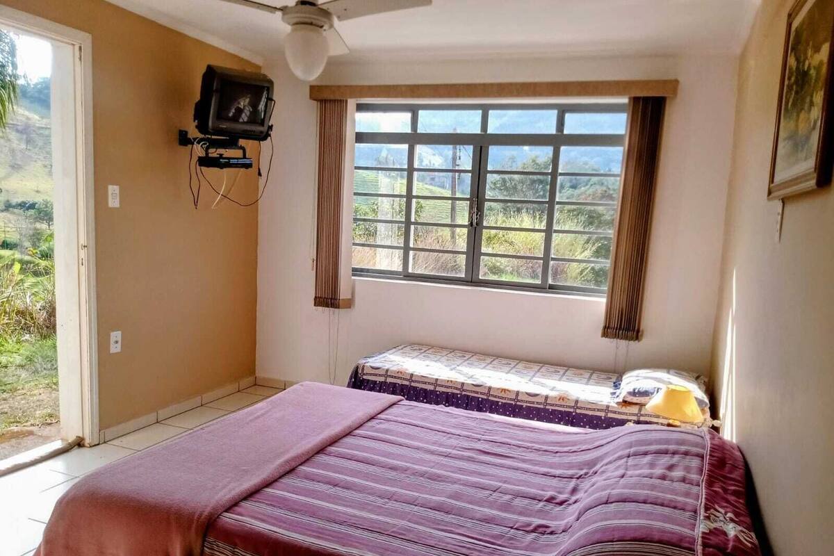 Ein Schlafzimmer mit einem Bett, einem Deckenventilator, einem Fenster mit Vorhängen und Blick ins Grüne.