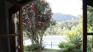 A view from a window overlooking a swimming pool, surrounded by lush greenery and mountains in the background. - Pousada Amanhecer na Mantiqueira (Águas de Lindóia)