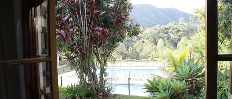 Vista desde una ventana con vista a una piscina, rodeada de exuberante vegetación y montañas al fondo.