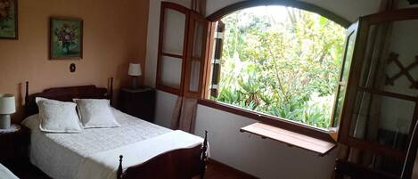 A bedroom with a wooden bed, a nightstand, a lamp, a window with a view of greenery, and a ceiling fan.