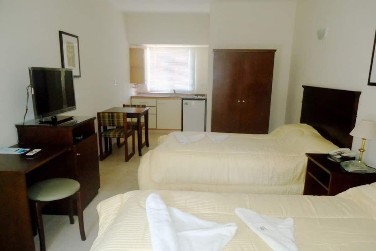 Habitación de hotel con dos camas, un escritorio con televisor, una silla y una pequeña kitchenette.