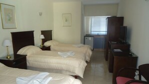Habitación de hotel con tres camas, un escritorio, una silla, un televisor y una ventana con persianas.