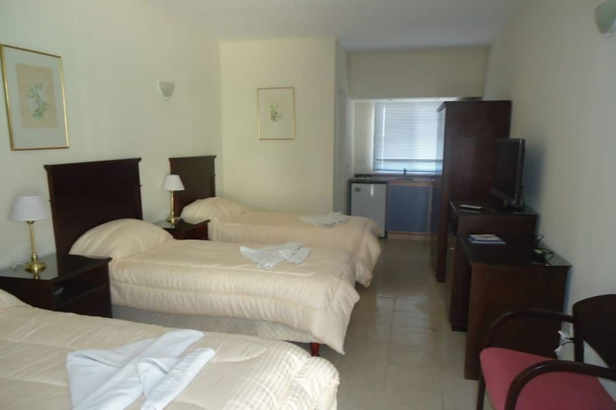 Habitación de hotel con tres camas, un escritorio, una silla, un televisor y una ventana con persianas.