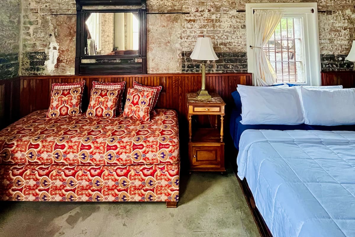 Una habitación con pared de ladrillo, cama con colcha azul, cabecera de madera, mesita de noche con lámpara y espejo en la pared.