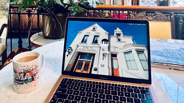 Um laptop sobre uma mesa com uma xícara de café, uma planta grande e vista de um edifício pela janela.