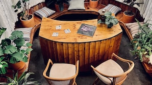 Uma mesa de madeira redonda e aconchegante rodeada por cadeiras estofadas, com plantas em vasos e um livro em cima.