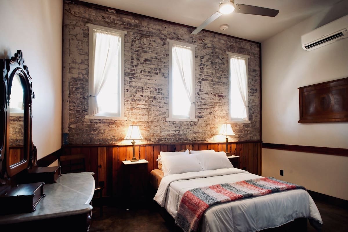 Un dormitorio con pared de ladrillo, dos ventanas con cortinas, una cama con edredón blanco, dos mesitas de noche con lámparas y un aparador de madera.