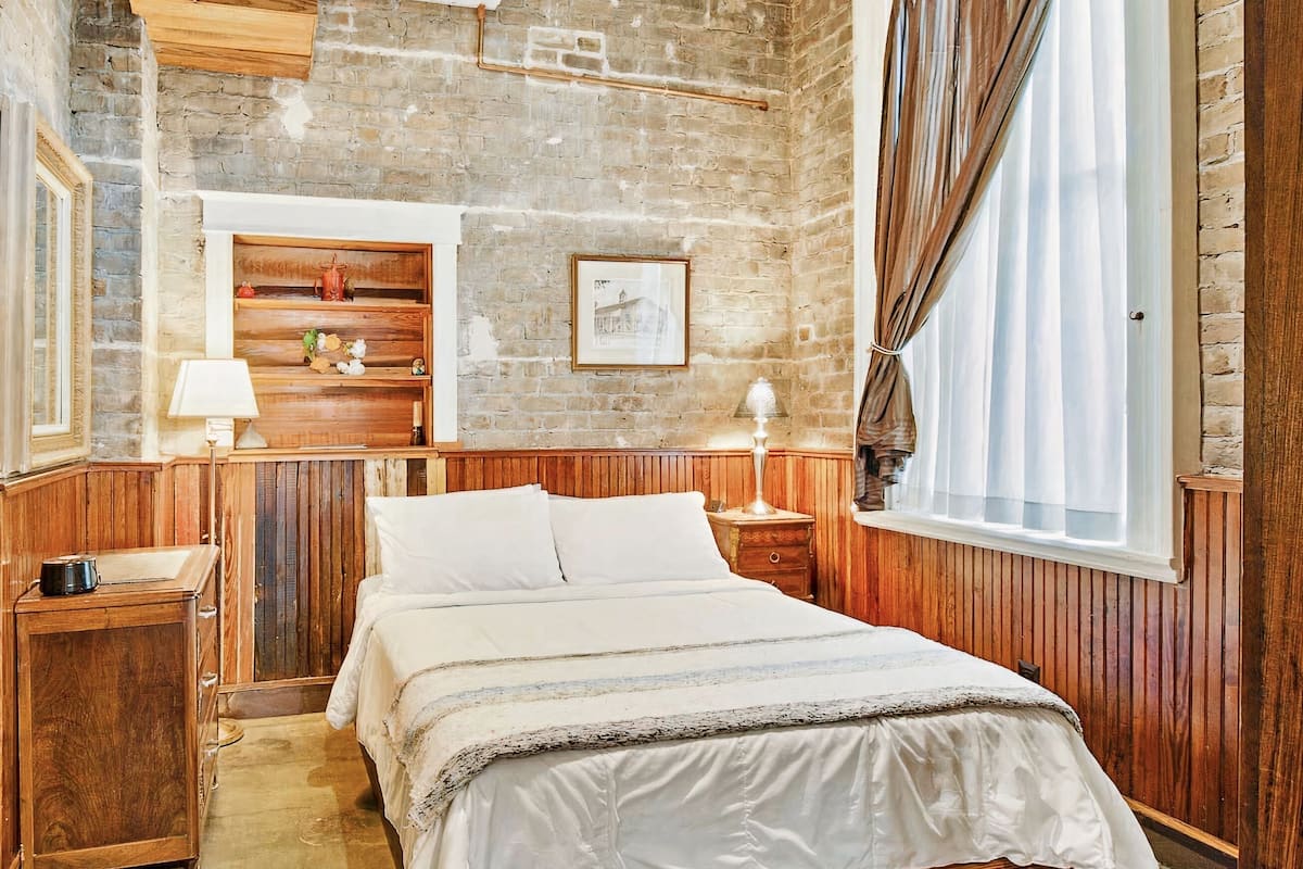 Un dormitorio con pared de ladrillo, paneles de madera, una cama con ropa blanca, una mesita de noche con lámpara y una ventana con cortinas.