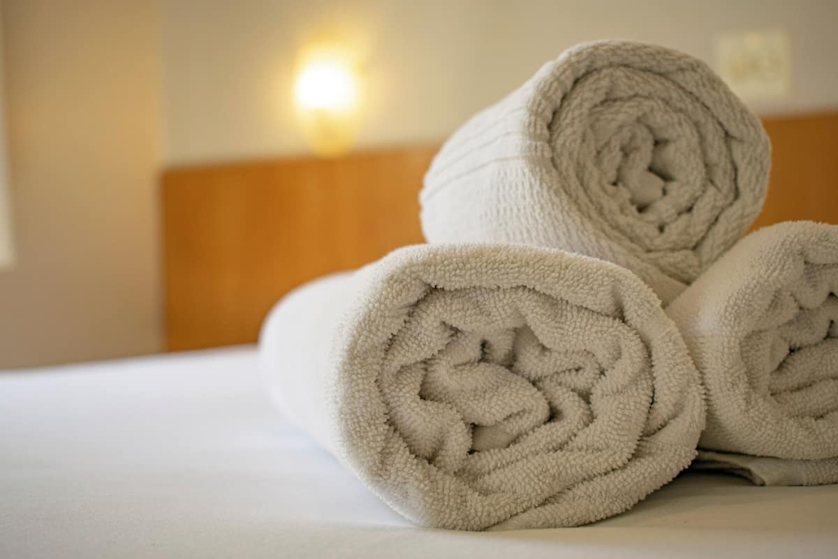 A neatly folded white towel on a bed with a wooden headboard and a wall-mounted light fixture in the background.