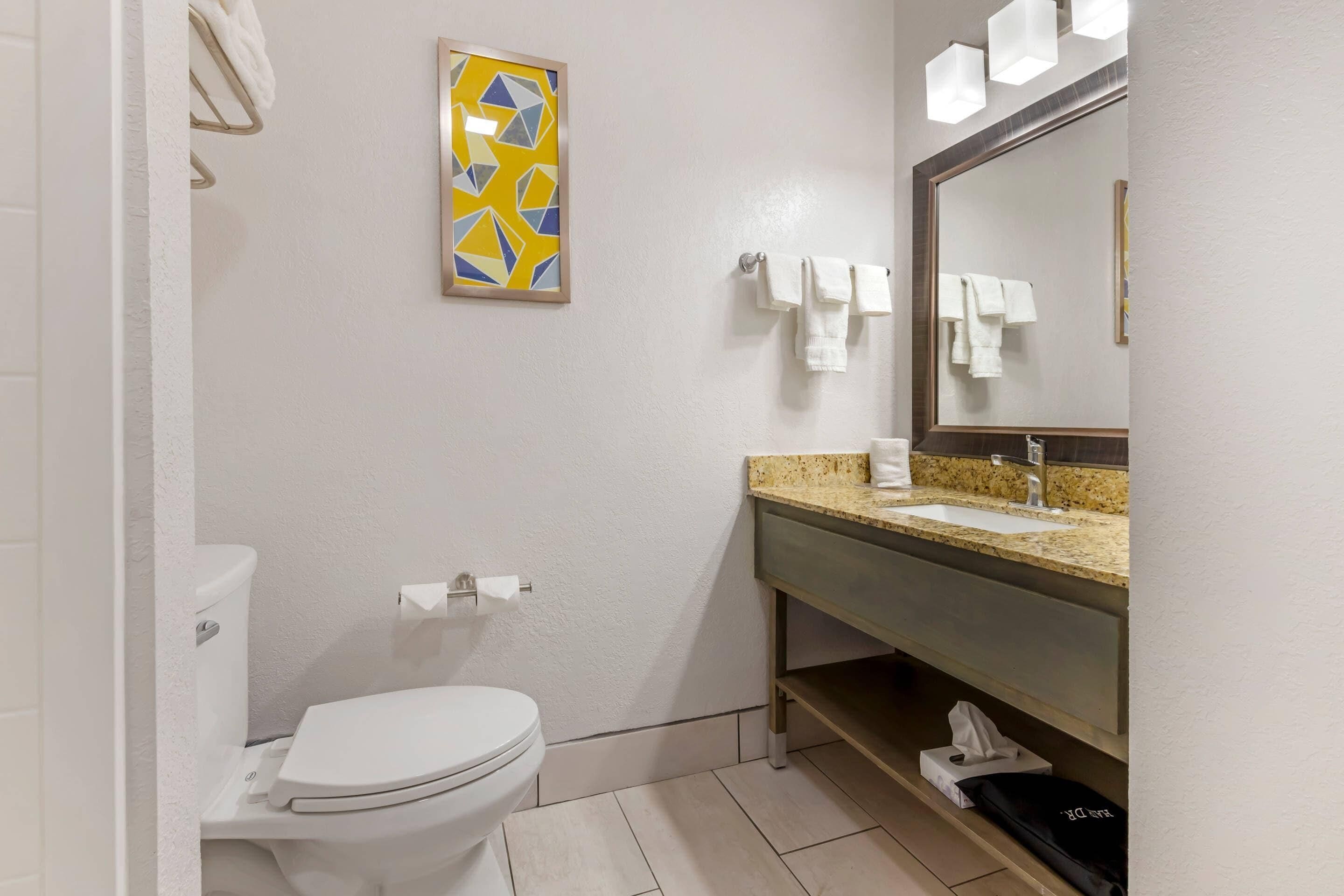 Un baño moderno con inodoro, lavamanos y espejo. Hay un perchero con toallas y un cuadro abstracto enmarcado en la pared.