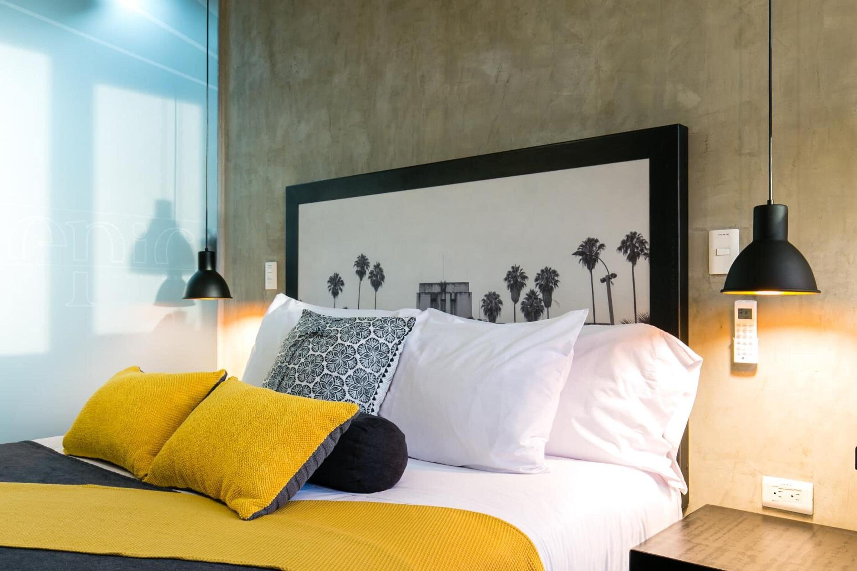 Una cama bien hecha con ropa de cama blanca y amarilla, un cuadro en la pared y dos lámparas colgantes negras.
