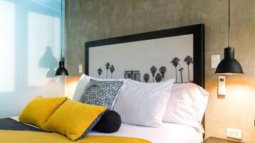 Una cama bien hecha con ropa de cama blanca y amarilla, un cuadro en la pared y dos lámparas colgantes negras.