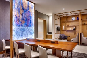Un salón de conferencias con una mesa de madera, sillas blancas, un gran mapa abstracto en la pared y un televisor en un estante.