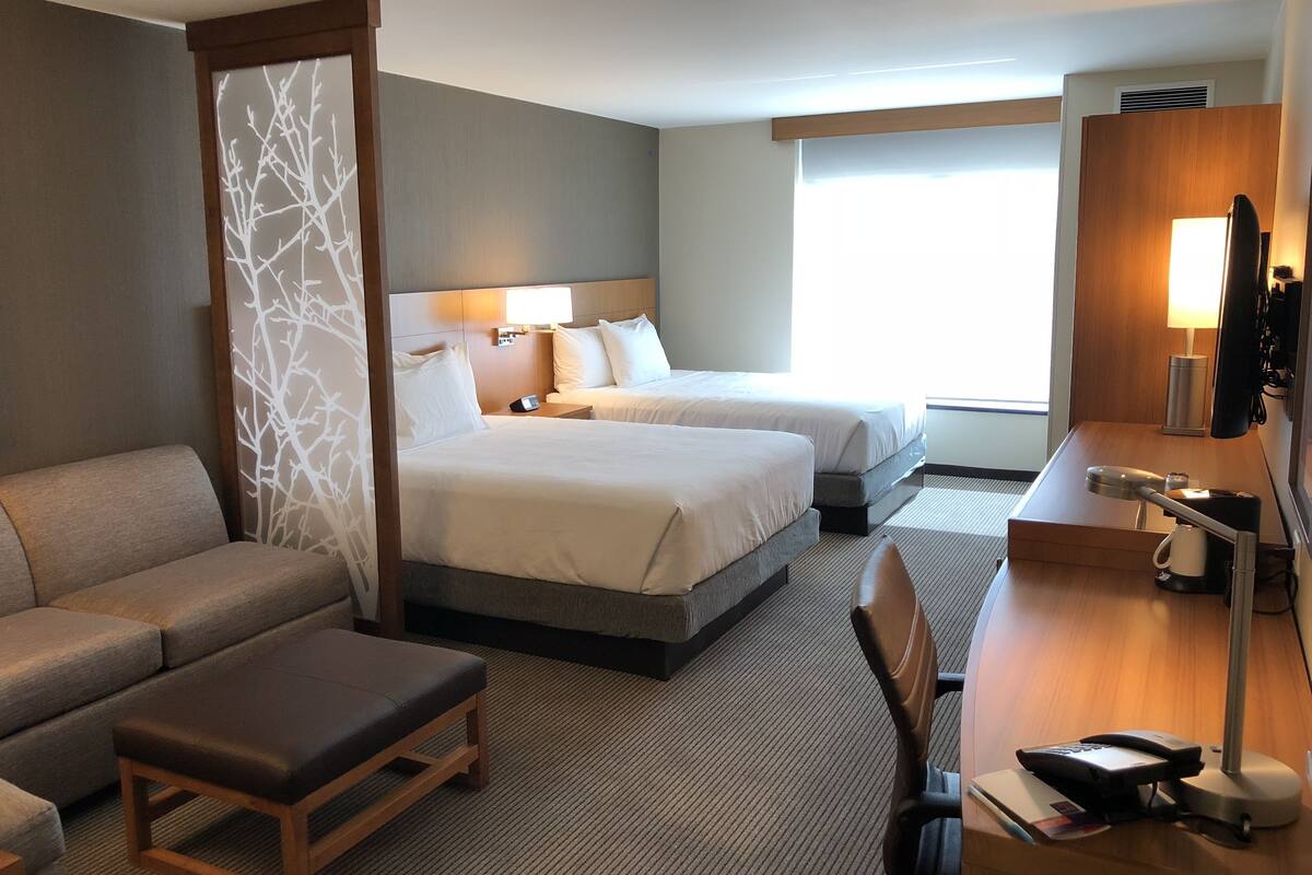A hotel room with two beds, a sofa, a desk with a phone, and a large window.