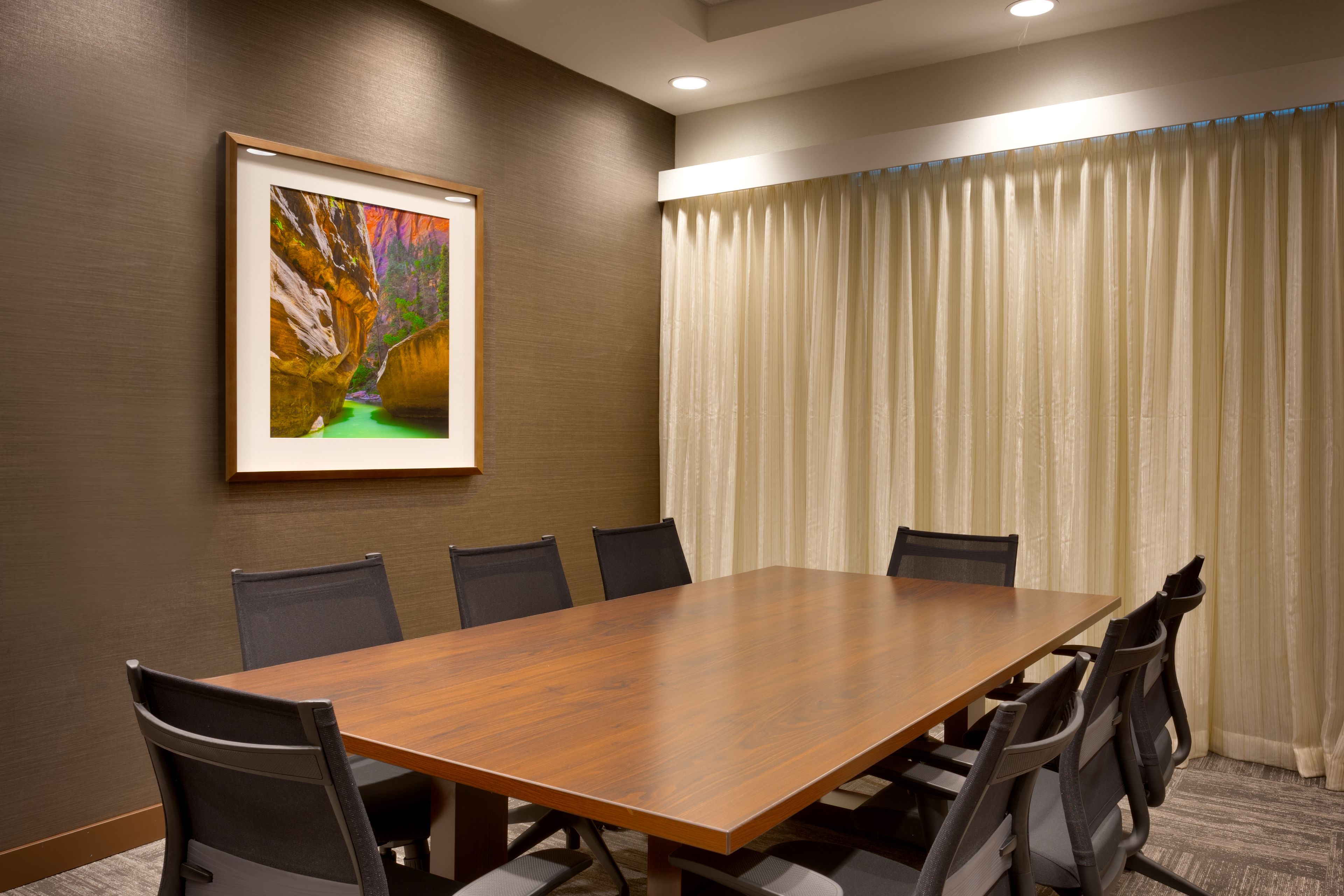 A conference room with a wooden table, six chairs, a framed picture on the wall, and a window with curtains.