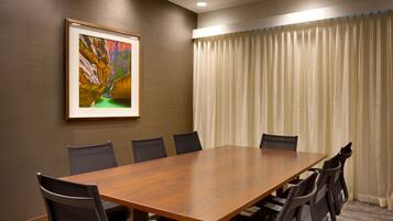 A conference room with a wooden table, six chairs, a framed picture on the wall, and a window with curtains.