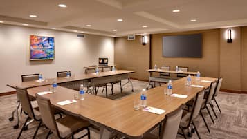 Un salón de conferencias con varias mesas y sillas, equipado con botellas de agua y vasos, una pantalla grande y un cuadro en la pared.