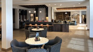 Een moderne hotellobby met een bar in het midden, zitgedeeltes en een keuken op de achtergrond.