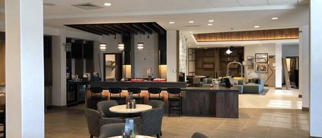 A modern hotel lobby with a central bar area, seating arrangements, and a kitchen in the background.