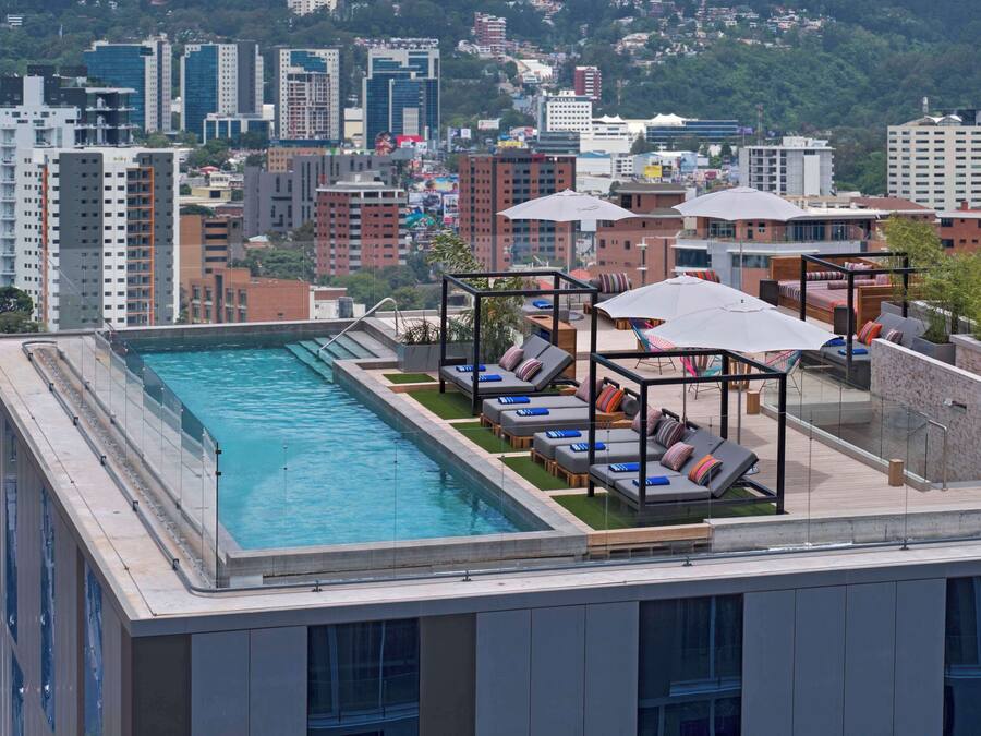 A rooftop pool area with sun loungers and umbrellas, surrounded by high-rise buildings.