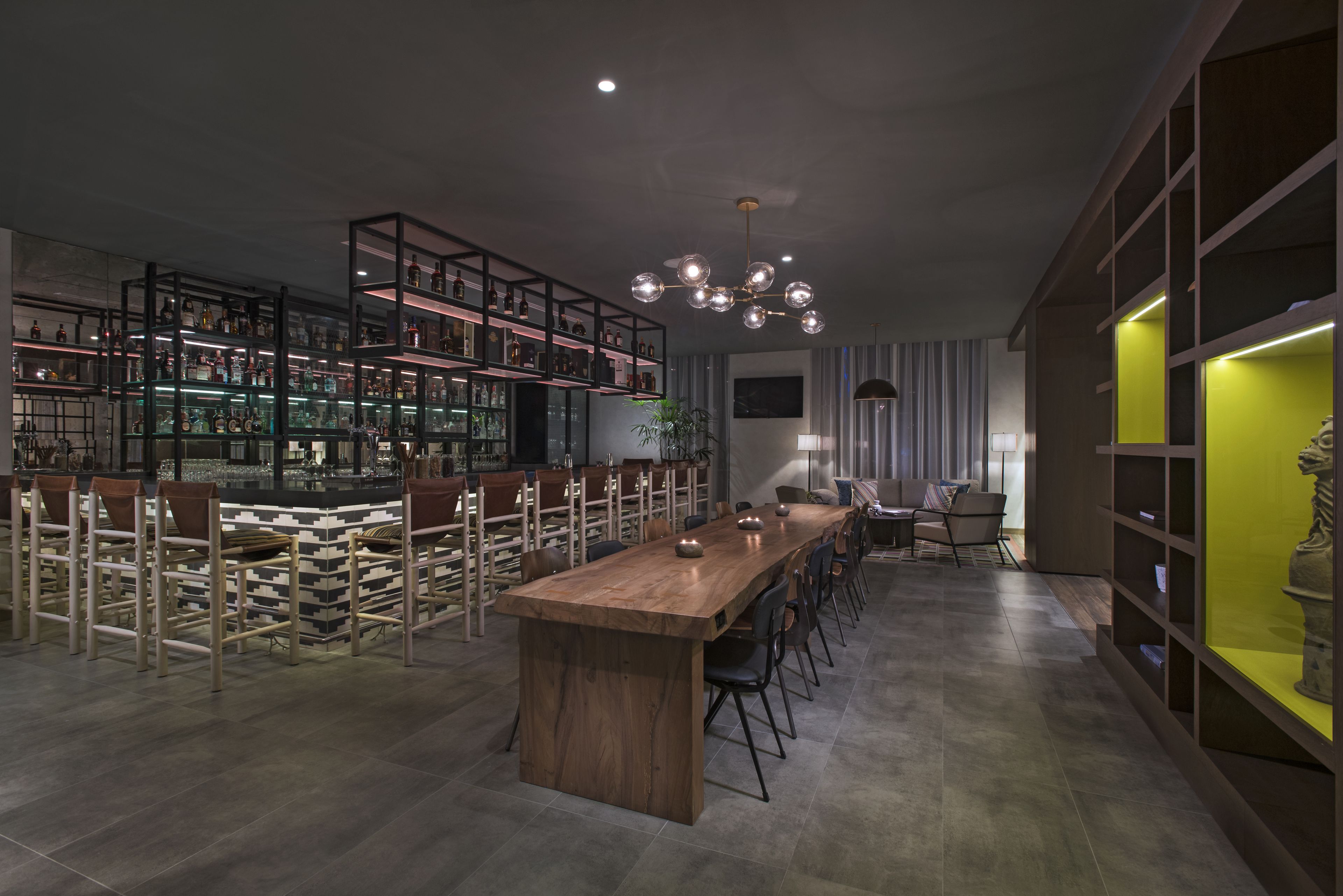 A modern bar area with a long wooden bar, multiple shelves with bottles, and a seating area with chairs and a table.