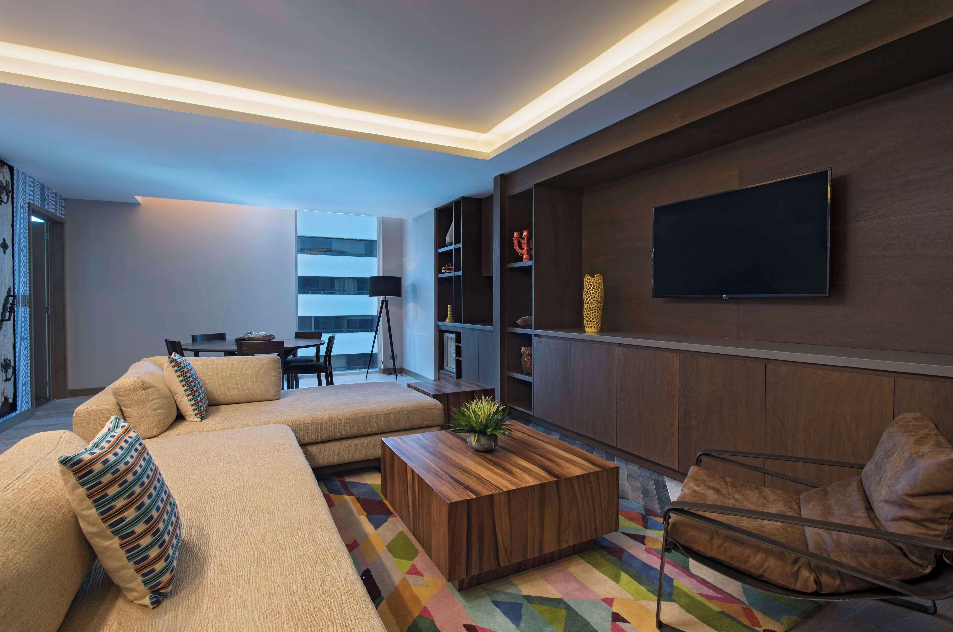 Una sala moderna con un sofá modular, una mesa de centro de madera, una alfombra colorida, un sillón de cuero y estanterías empotradas con un televisor de pantalla plana.