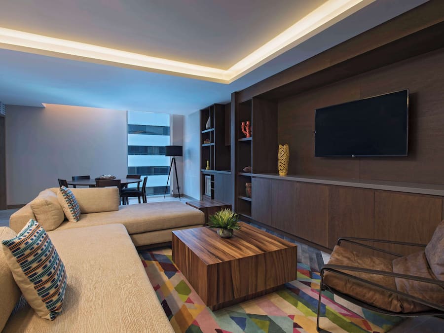 Una sala moderna con un sofá modular, una mesa de centro de madera, una alfombra colorida, un sillón de cuero y estanterías empotradas con un televisor de pantalla plana.